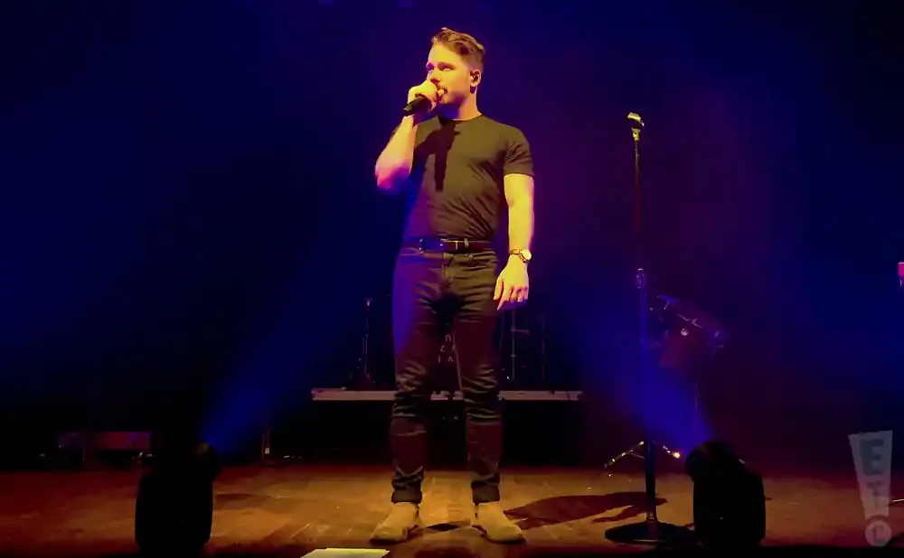 A man in a black t-shirt and jeans sings into a microphone on a stage with blue lighting.