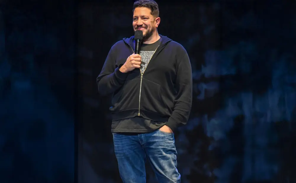 A man in a black hoodie and jeans smiles while holding a microphone on stage.