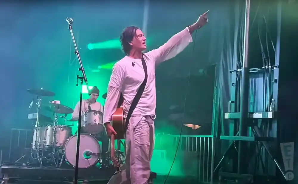 A male musician in a white shirt and pants plays guitar on stage, pointing his finger to the right, with a drummer in the background.