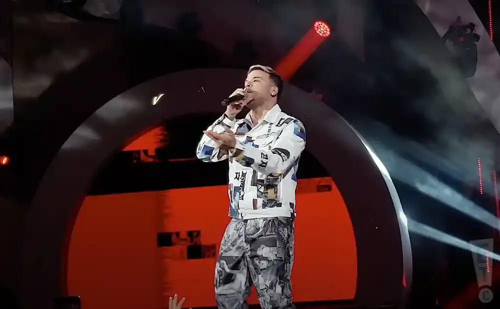 A man in a patterned shirt and pants sings into a microphone on a stage with red and black accents.