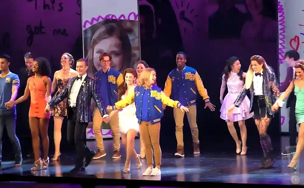 A musical theater cast, some in letterman jackets, takes a bow on stage.