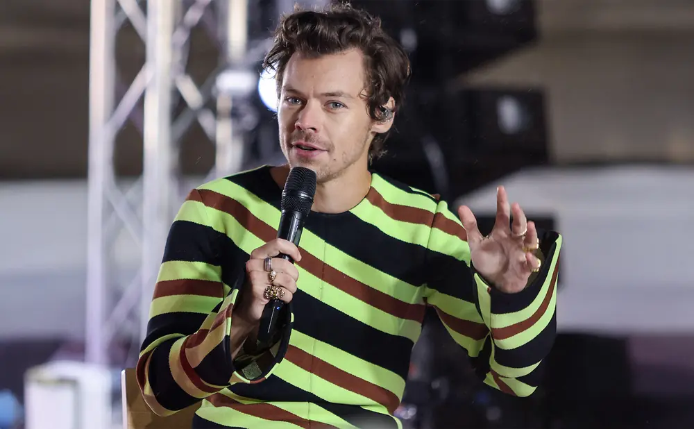 Harry Styles, wearing a striped shirt, speaks into a microphone and gestures with his hand.