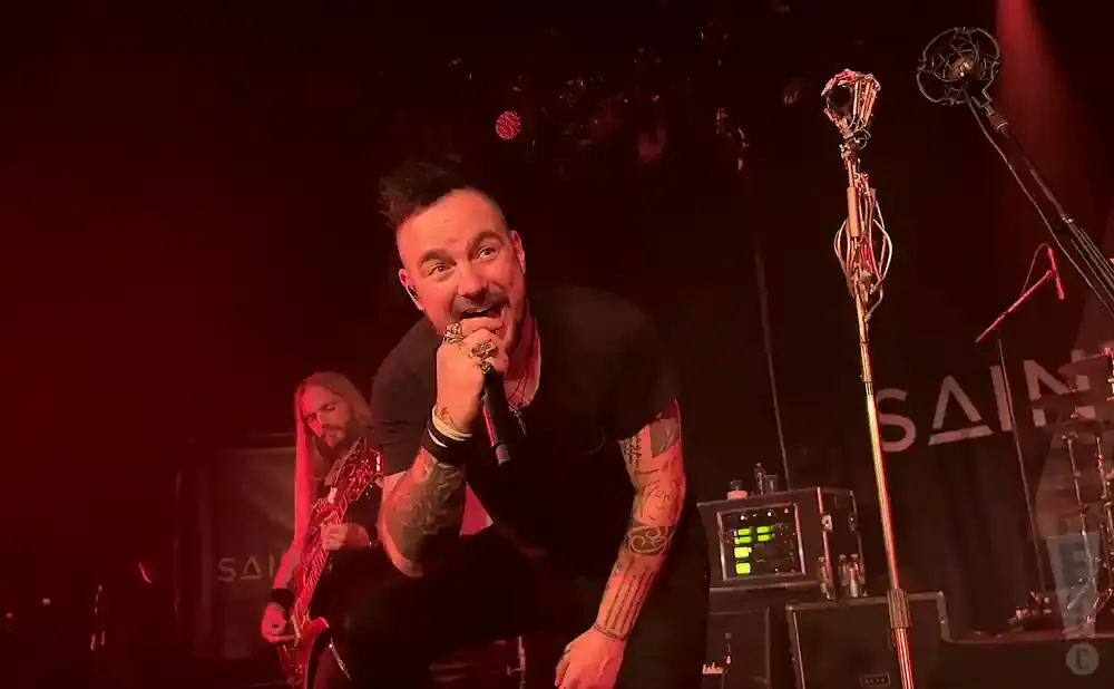 A male singer with tattoos and spiky hair sings into a microphone on stage, with a guitarist behind him.