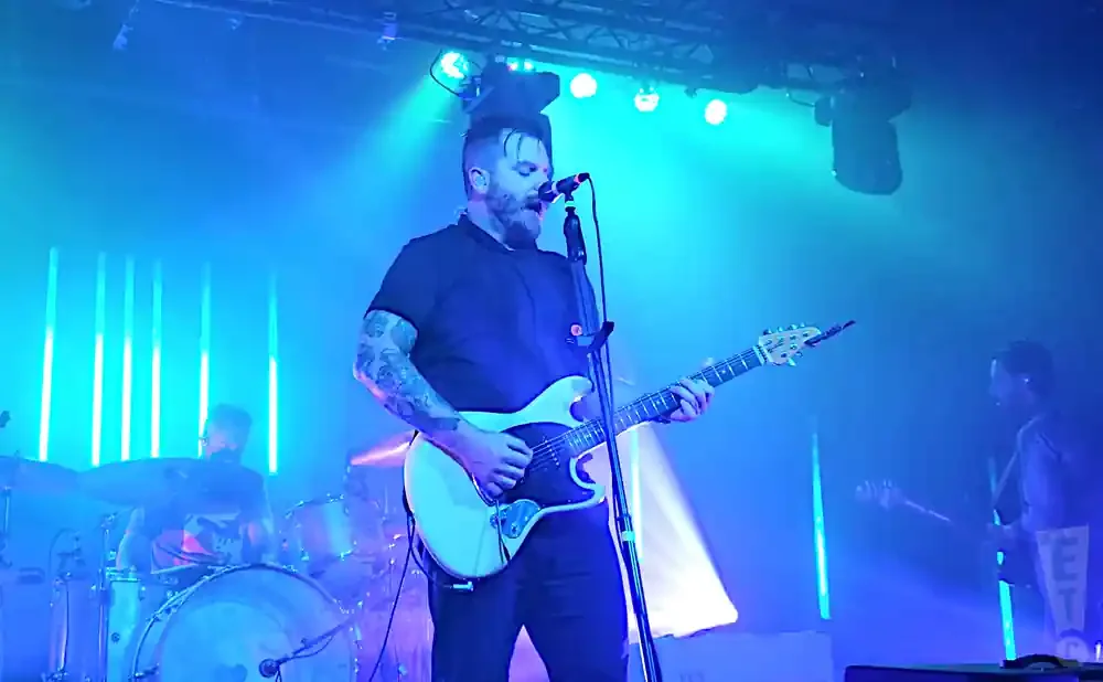A male musician sings and plays a white electric guitar on a stage lit by blue lights.
