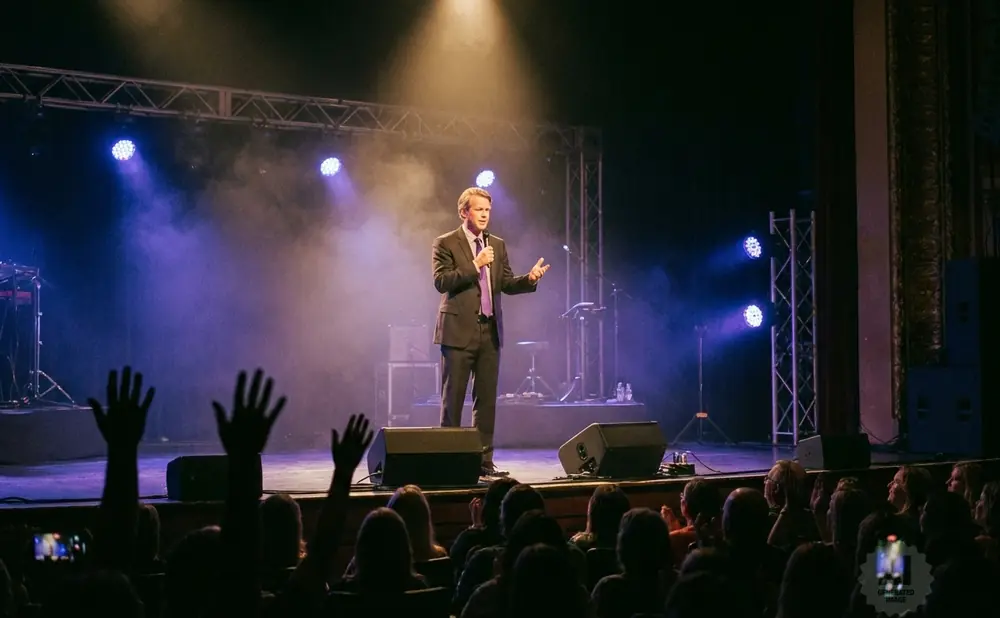 A man in a suit speaks into a microphone on stage, with a projection screen behind him and an audience in the foreground.