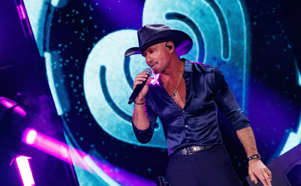 Man in a black cowboy hat and open-collared blue shirt sings into a microphone on stage with a blue and purple LED screen behind him.