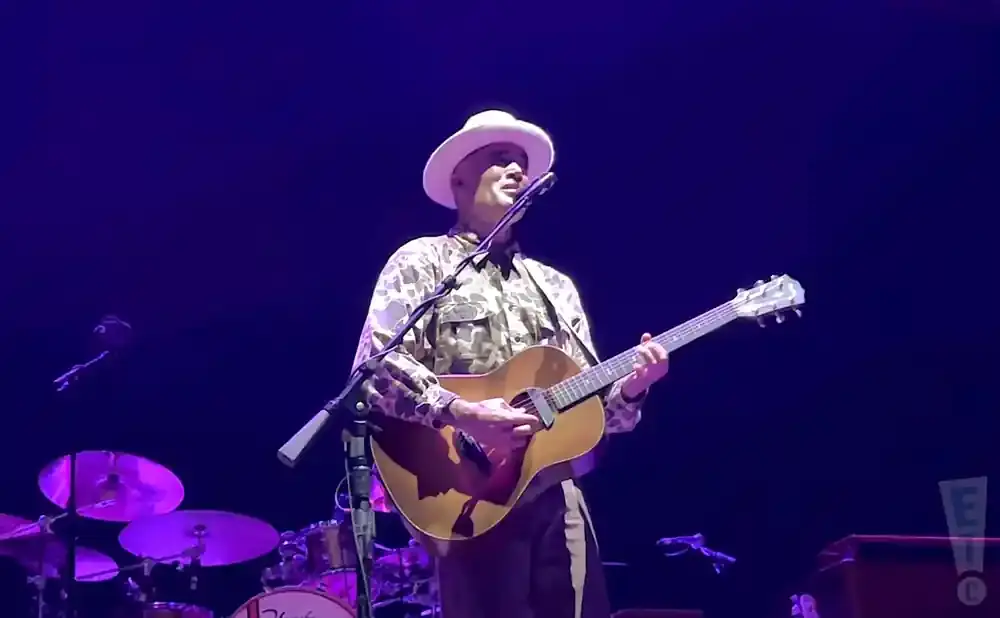 A man in a white hat and camouflage shirt plays an acoustic guitar on stage.