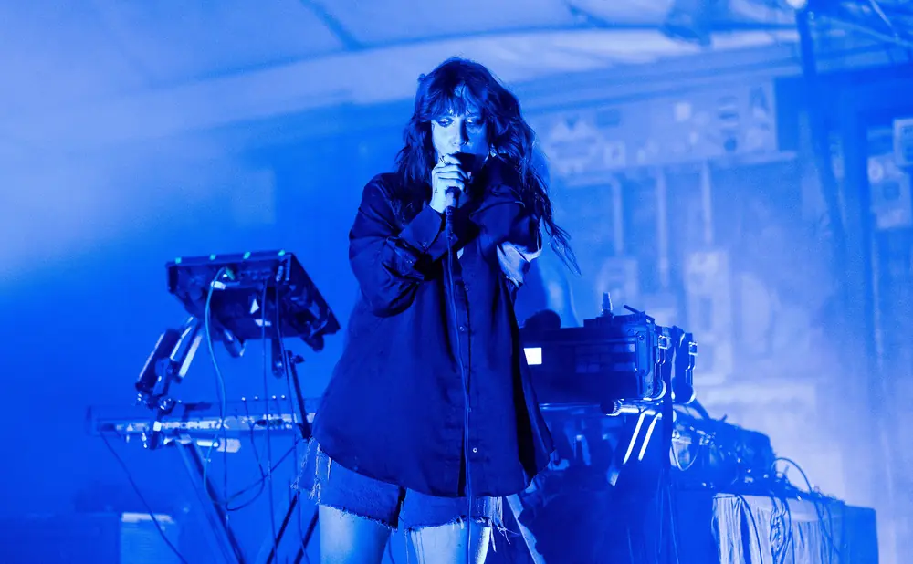Singer in a dark shirt and denim shorts, holding a microphone with a blue stage light.
