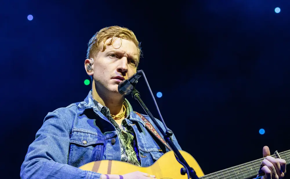Musician with blonde hair plays an acoustic guitar and sings into a microphone while wearing a denim jacket.
