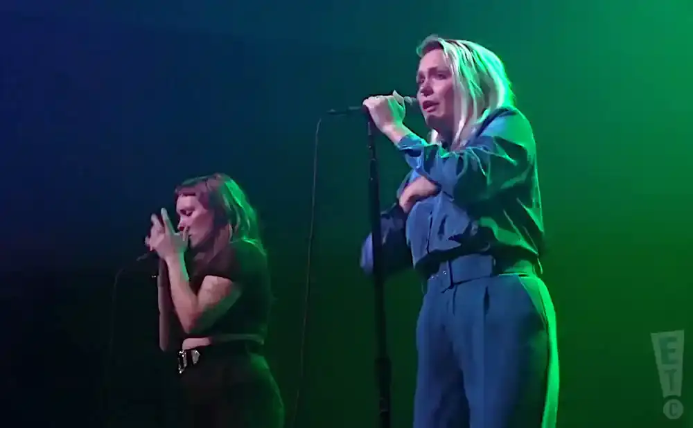 Two women sing into microphones on a dimly lit stage with green and blue lights.