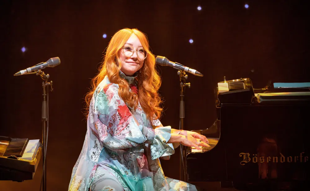 A redheaded woman in glasses plays a Bösendorfer piano on stage.