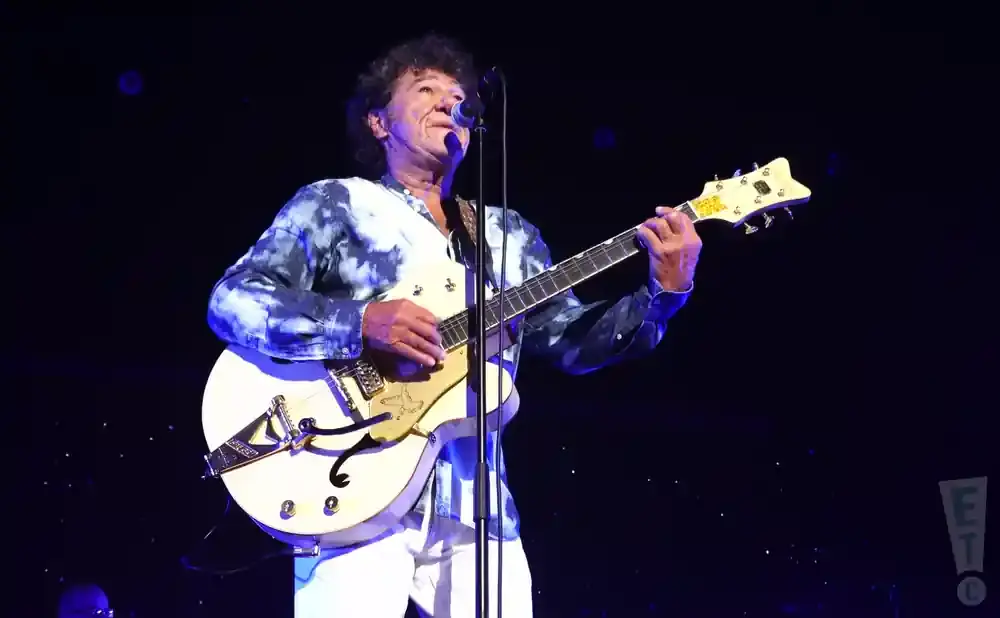 Musician with curly hair plays a white electric guitar on stage, wearing a tie-dye shirt and white pants.