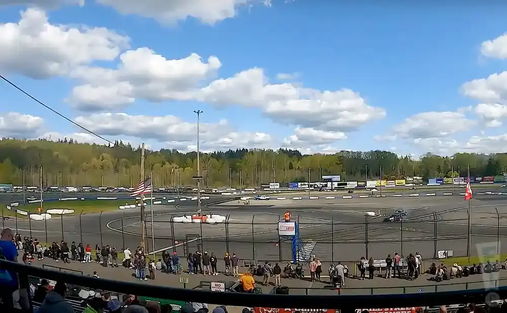 A race car speeds around a track, with spectators in the foreground and a forest backdrop under a cloudy sky.