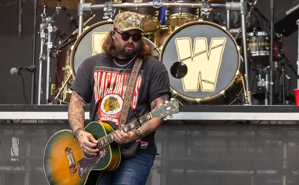 Musician playing an acoustic guitar on stage with drums in the background.