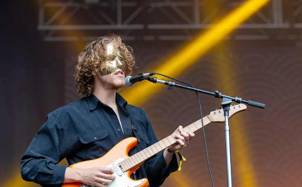 A masked guitarist plays an orange electric guitar on stage under yellow lights.