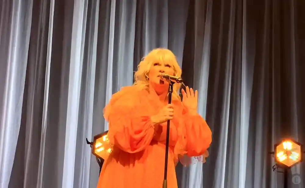Singer with blonde hair in an orange dress and fur stole sings into a microphone on stage with white curtains and stage lights.