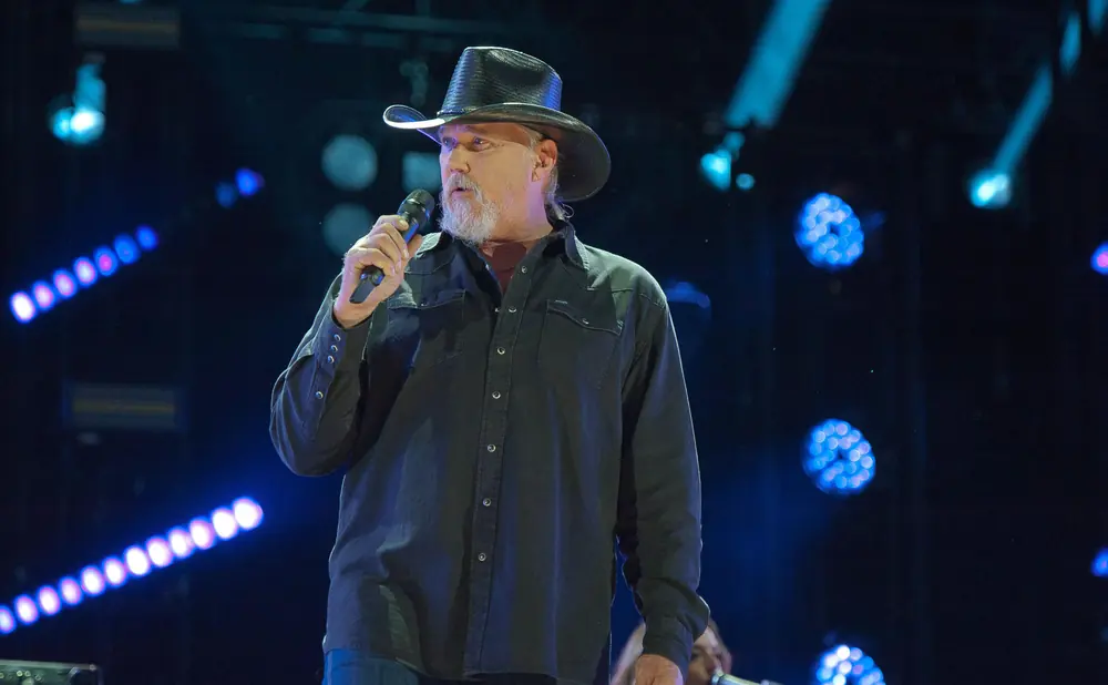 Man in a cowboy hat and black shirt sings into a microphone on a stage with blue lights.