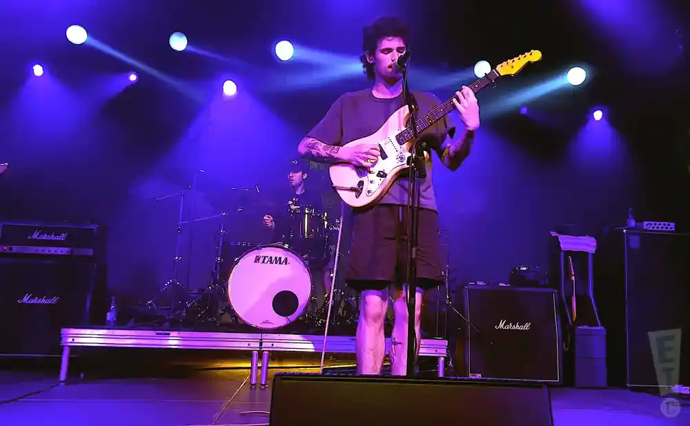 A male guitarist plays on stage with purple lighting and a drummer in the background.