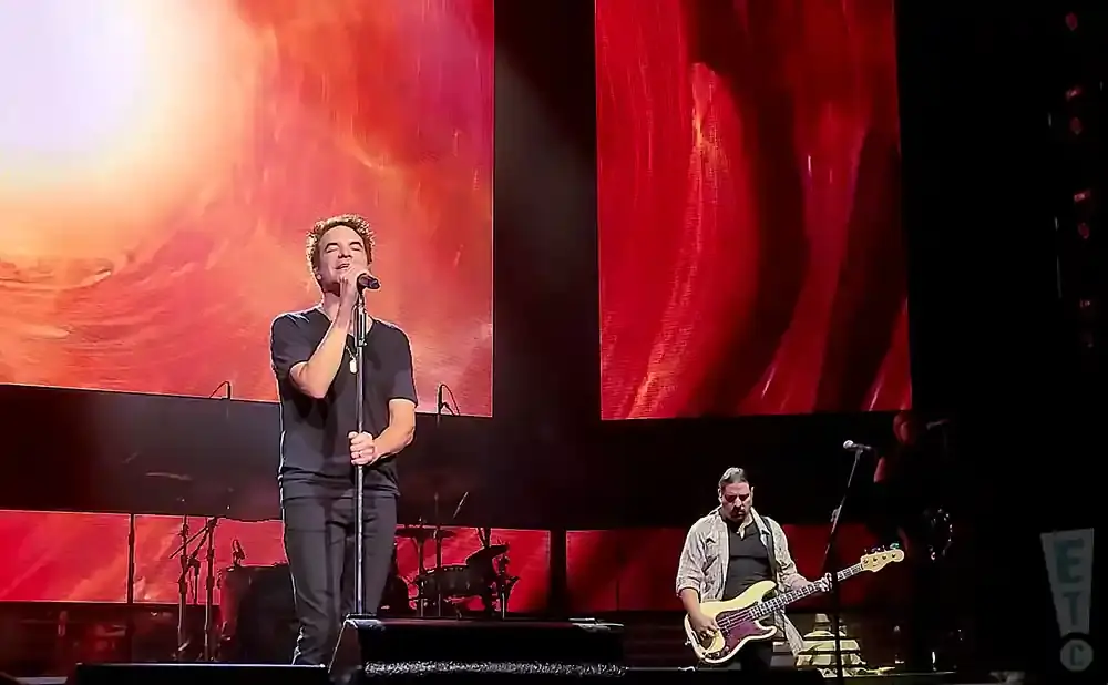 A man sings into a microphone on stage, while another plays bass guitar. Red and orange screens are behind them.