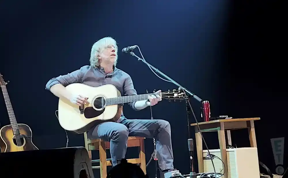 A musician with blonde hair plays an acoustic guitar while sitting on a stool on stage.