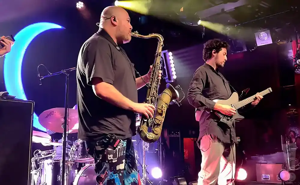 A bald man plays a saxophone onstage in front of a blue crescent moon, while a man in a black shirt plays guitar.