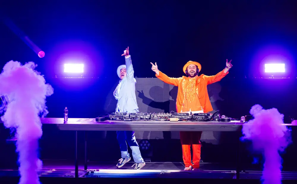 Two DJs in bright outfits enthusiastically perform on stage with purple smoke and vibrant blue lighting.
