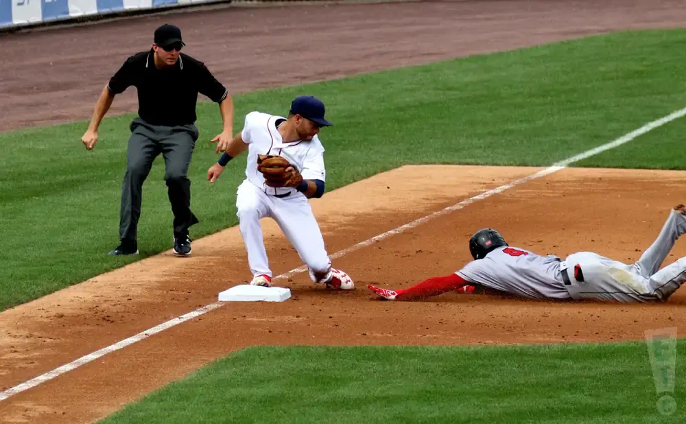 Baseball players slide into home base as an umpire watches.