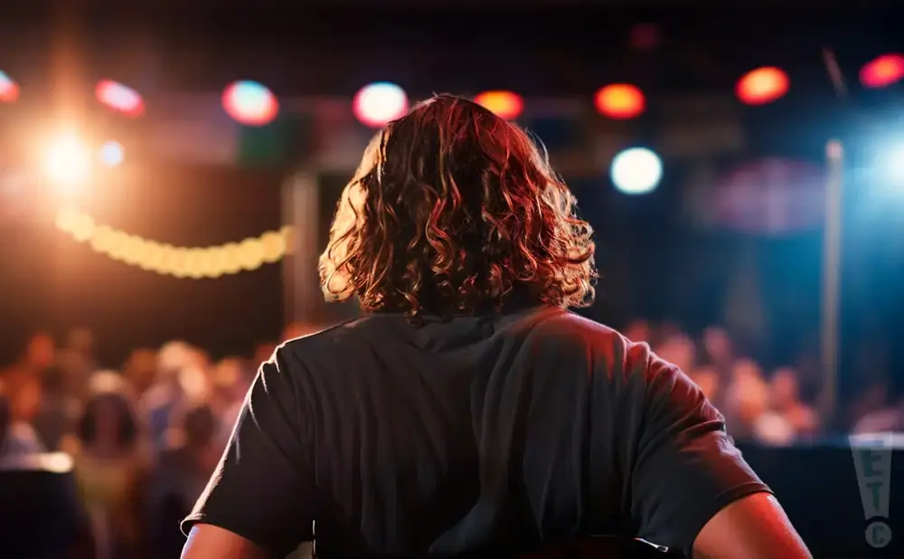 Man with curly hair facing away from camera on stage, with blurred audience and colorful lights in the background.
