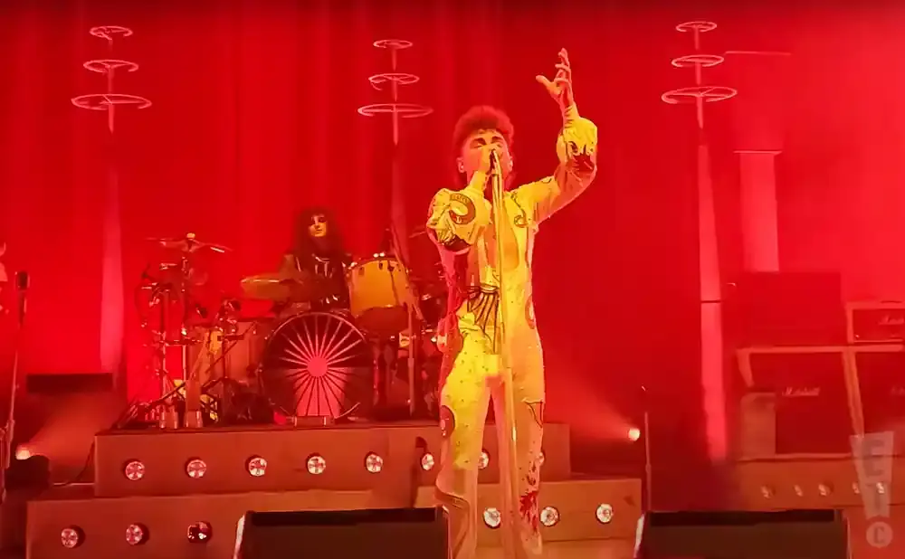 A performer in a yellow patterned jumpsuit sings on stage with a drummer in the background.