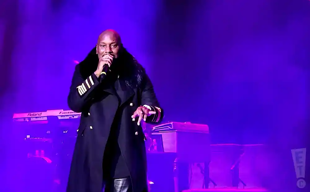 A bald man in a black coat with fur trim sings into a microphone on a stage with purple lighting and keyboards.