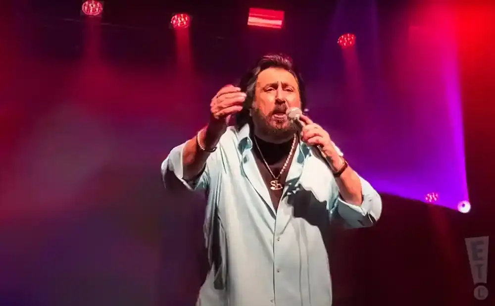 Singer wearing a light blue shirt and gold necklace performs on a stage with red and purple lights.