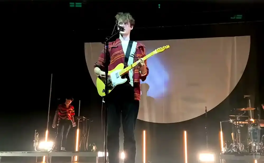 A man in a striped shirt plays a yellow electric guitar on stage.