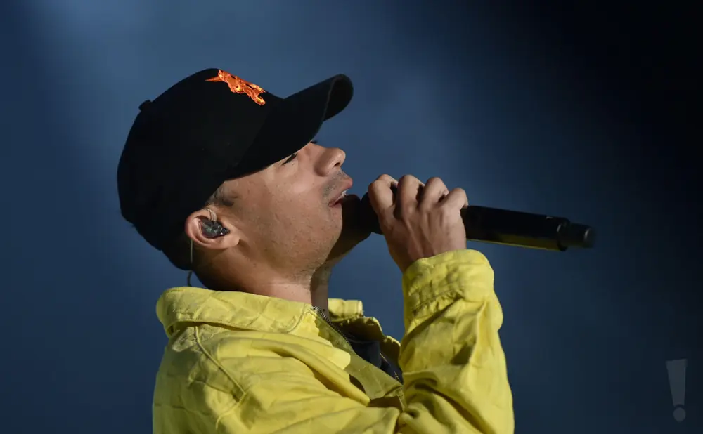 A man in a yellow jacket and black cap sings into a microphone with an earbud in his ear.