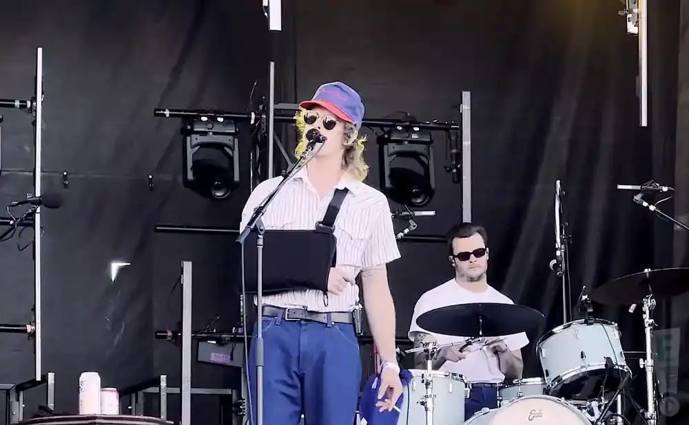 A male singer in a striped shirt and blue cap sings into a microphone while a drummer plays behind him on stage.
