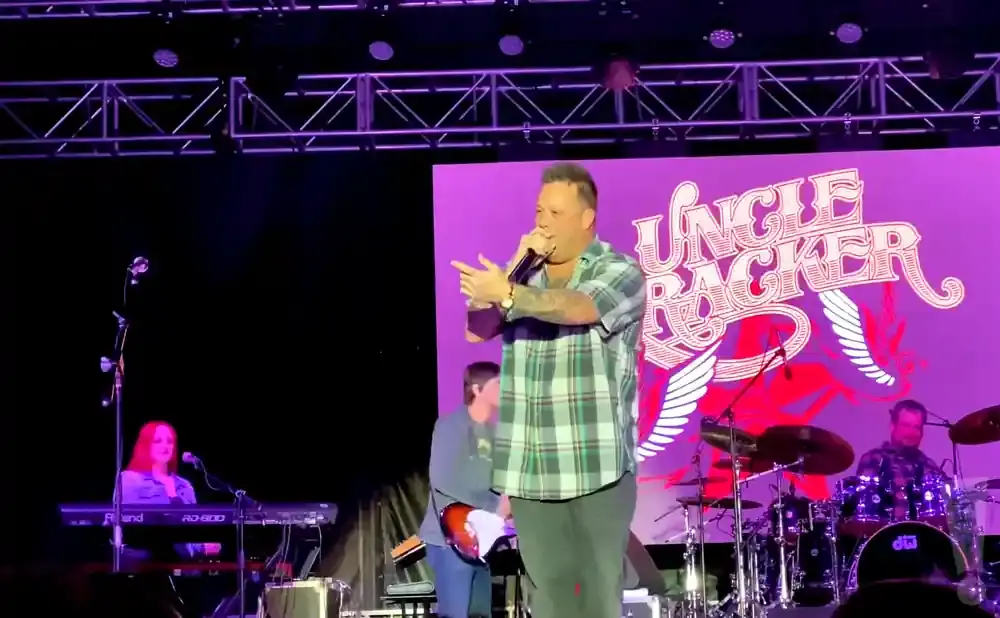 A male singer in a plaid shirt performs on stage with a band behind him.