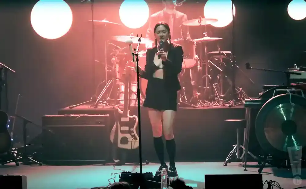 A woman in a black outfit sings into a microphone on a stage with a drum set and guitars.