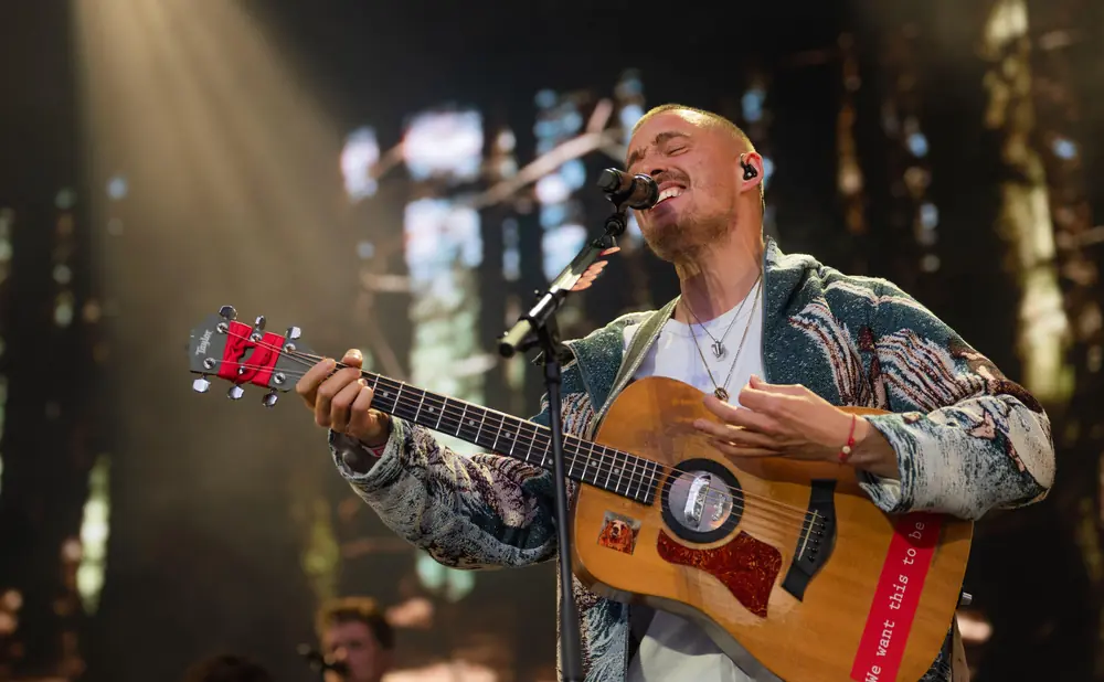 A man sings and plays a guitar on stage, wearing a patterned jacket and necklaces.