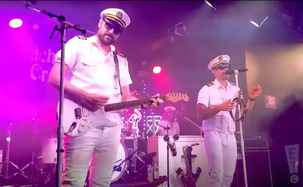 Musicians in white captain's uniforms perform on stage under purple lights, one playing guitar, the other a tambourine.