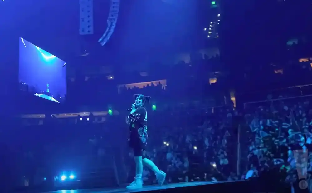 Billie Eilish performs on a brightly lit stage at a concert, with a large audience in the background.