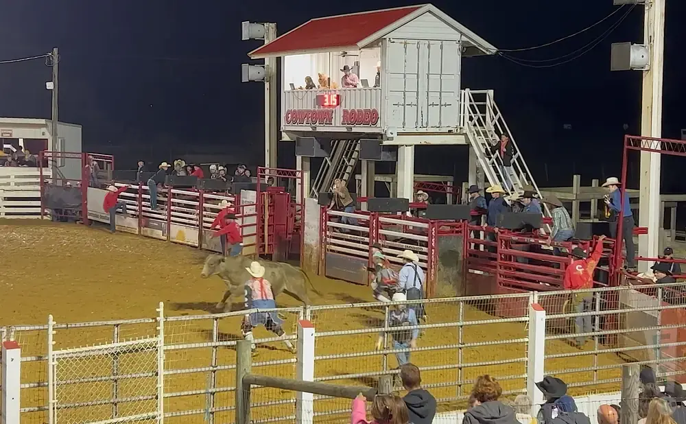Rodeo action at night: cowboys in red shirts and hats on arena floor with bull, crowd in stands, and announcer booth.
