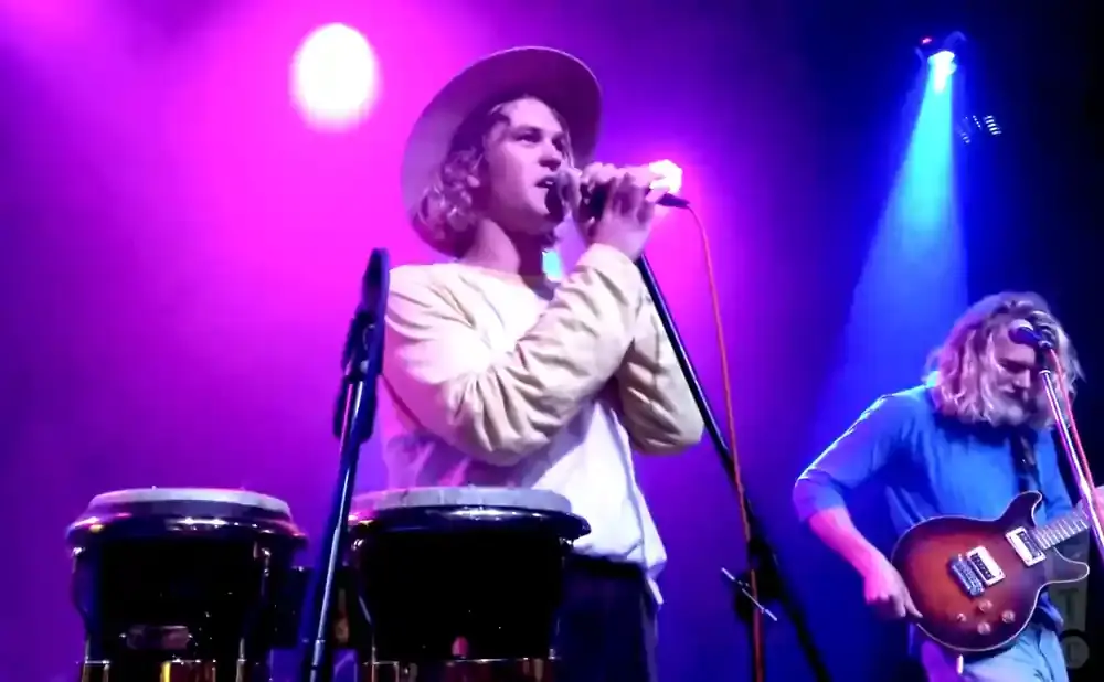A band performs on a stage with purple lighting; the singer wears a hat and tan shirt.