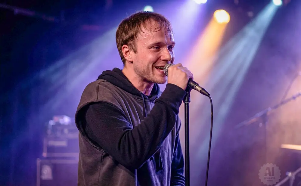 A male singer sweats as he performs into a microphone on a dimly lit stage.