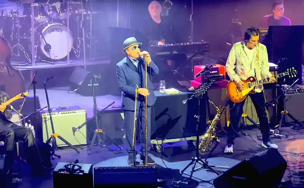 A male singer in a blue suit and hat plays the trombone on stage with a band.