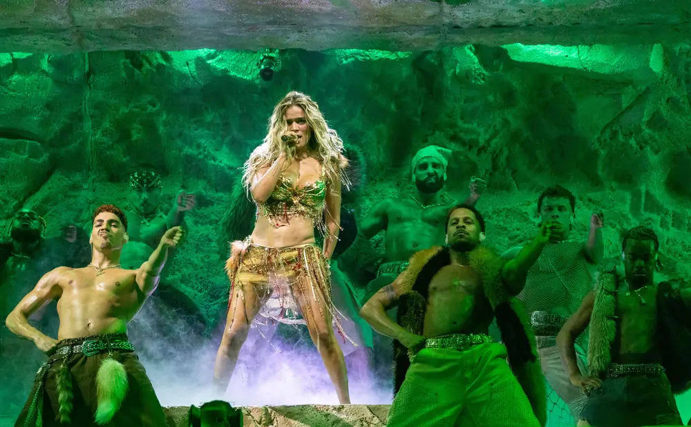 A female singer in a gold costume performs on stage with backup dancers in tribal attire, illuminated by green lights.