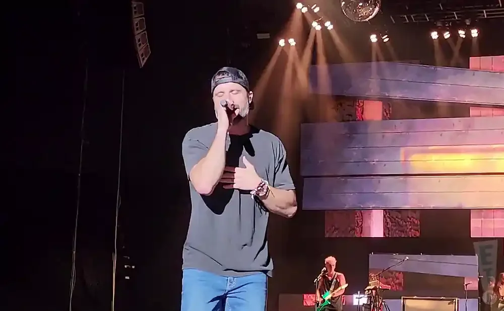 Singer in a gray t-shirt and jeans performs on stage, holding a microphone and touching his chest.