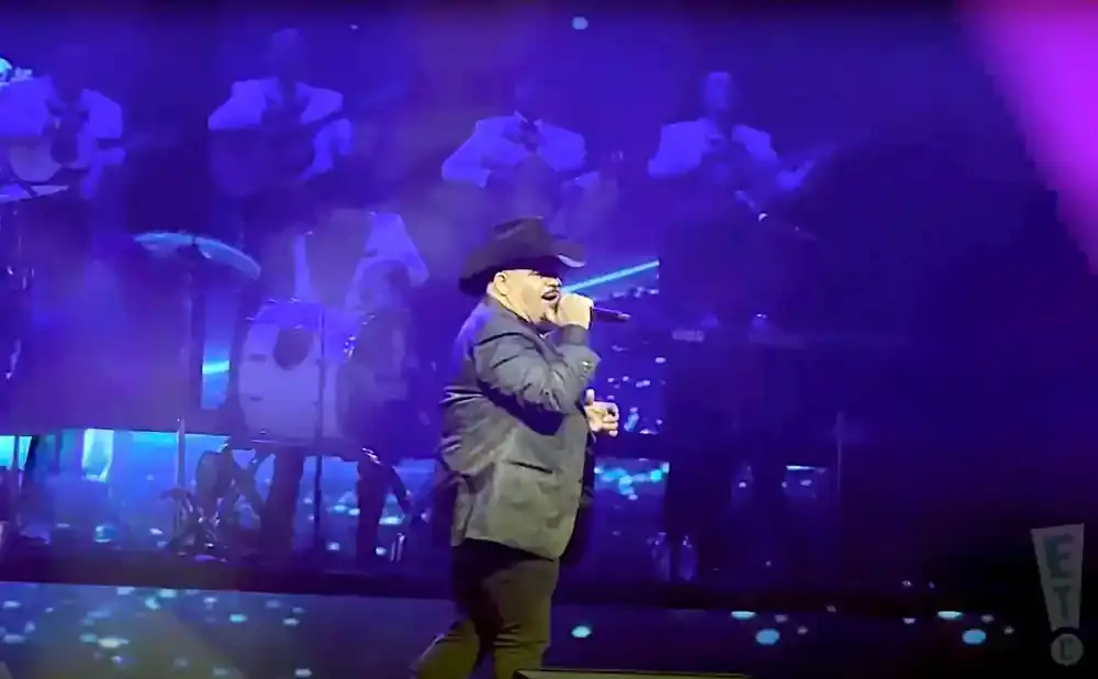 A man in a cowboy hat sings into a microphone on a stage with a band behind him.