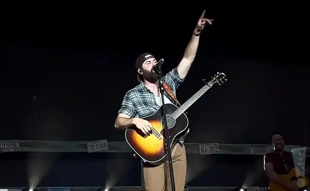 A man in a plaid shirt and baseball cap plays guitar and sings on stage, raising his hand.