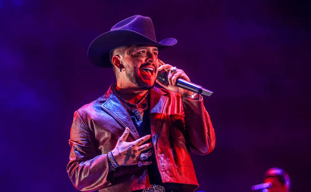 A man in a cowboy hat and leather jacket sings into a microphone under purple and red lights.