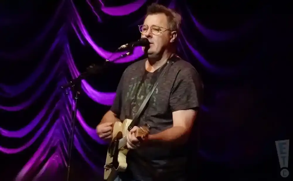 Man with glasses and a gray shirt plays a cream-colored guitar and sings into a microphone on a stage with purple lighting.