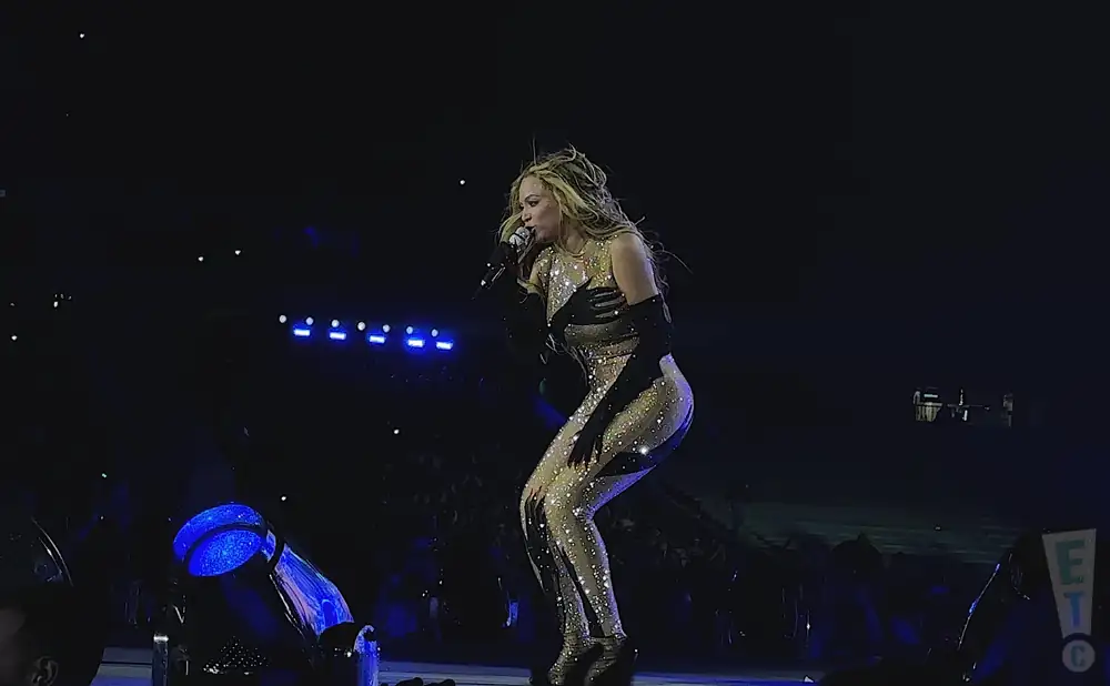 A singer in a sparkly nude and black outfit performs on stage with blue lights in the background.
